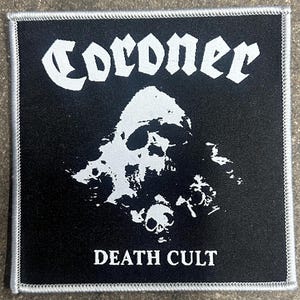 May include: Square black patch with white embroidered border. The patch features the word "Coroner" in gothic font above a white skull graphic. Below the skull, the words "Death Cult" are printed in white.