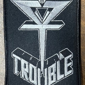 May include: A black embroidered patch with a white stylized logo of a triangle with a vertical line through it and the word "TROUBLE" below it.