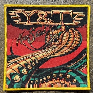 May include: A yellow and black embroidered patch with the band name "Y&T" and the album title "Mean Streak" in a stylized font. The background features a red and gold illustration of a mechanical arm.