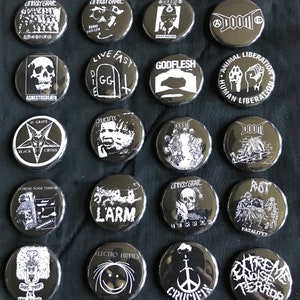 May include: A collection of 18 black and white button pins with various designs. The pins feature band names, logos, and slogans. Some of the designs include "Unholy Grave", "Godflesh", "Doom", "Crucifix", "Larm", "Animal Liberation", "Human Liberation", "Asbestosdeath", "Extreme Noise Terror", "Electro Hippies", and "Fatality".