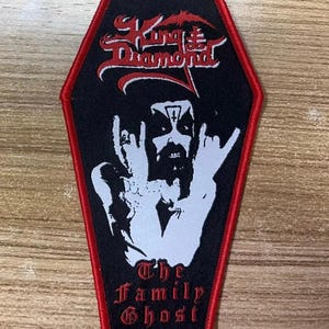 May include: Black and red embroidered patch with a coffin shape. The patch features the text "King Diamond" in red and "The Family Ghost" in white. A white image of a person with horns and a pentagram on their forehead is in the center of the patch.