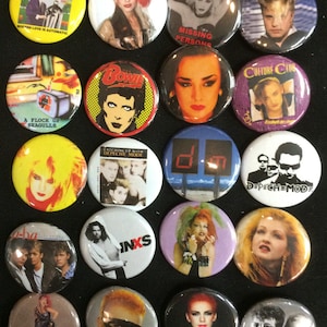 May include: A collection of round, colorful pinback buttons featuring images of 1980s musicians and bands. The buttons display portraits of artists like David Bowie, Cyndi Lauper, and members of Depeche Mode, along with band logos and album art.