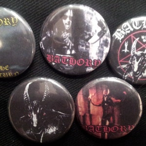 May include: A collection of five black Bathory band button pins. The pins feature images of the band members, the band's name, and other gothic imagery. The text "The Return" is on one pin.