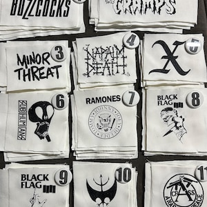 May include: A collection of eleven white fabric patches featuring black screen-printed band logos and graphics, including Buzzcocks, The Cramps, Minor Threat, Napalm Death, X, Subhumans, Ramones, Black Flag, Battalion of Saints, and Crass.