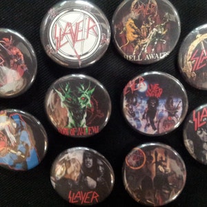 May include: Ten round metal pins with Slayer logos and album art. The pins feature images from albums such as "Reign in Blood", "Seasons in the Abyss", and "Hell Awaits".