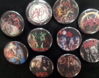 May include: Ten round metal pins with Slayer logos and album art. The pins feature images from albums such as "Reign in Blood", "Seasons in the Abyss", and "Hell Awaits".