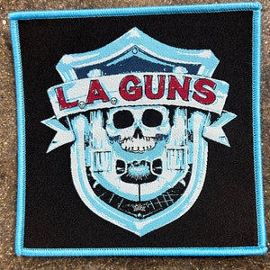May include: A blue and white embroidered patch with a skull and crossbones design. The text "LA GUNS" is in red letters above the skull.