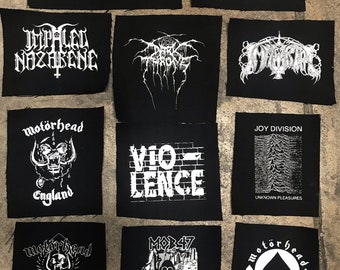 May include: A collection of black fabric patches with white text and graphics. The patches feature band logos and text, including "Immortal," "Motörhead," and "Joy Division." The patches are rectangular and have a raw edge.