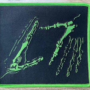May include: A black square patch with a neon green border and a design of two skeletal hands. The hands are also neon green and appear to be reaching out. The patch is likely for a jacket or bag.