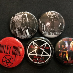 May include: Five round, black and white button pins with a band theme. The pins feature the band name "Motley Crue" and images of the band members. One pin has a pentagram with the text "Show at the Devil".