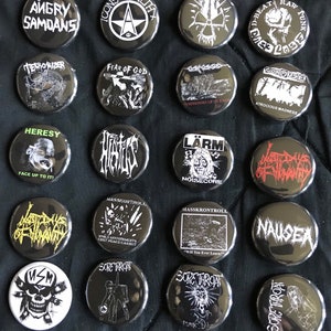 May include: A collection of 16 black and white round buttons with band names and logos. The buttons feature various punk rock and metal bands, including Angry Samoans, Terrorizer, Fear of God, Heresy, Lärm, Masskontroll, Sore Throat, and Nausea.