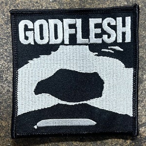 May include: Black and white embroidered patch with the word "GODFLESH" in white lettering. The patch features a stylized image of a face with a black eye and a white mouth.