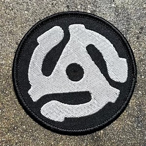 May include: A black and white embroidered patch with a stylized record player design. The patch is circular and has a black background with a white record player design.