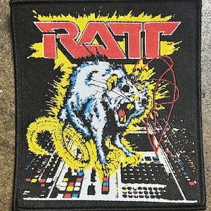 May include: A black and yellow embroidered patch with the word "RATT" in red letters and a white rat with red eyes and a yellow tail standing on a black and white soundboard.