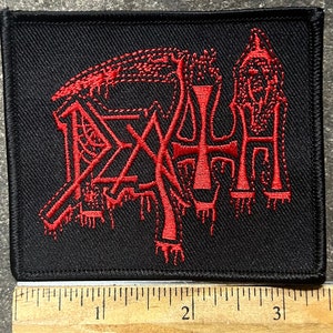 May include: Black embroidered patch featuring the word "DEATH" in a stylized, gothic font. The red lettering has a dripping effect, and the patch is rectangular. The patch is on a black background.