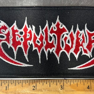 May include: Black rectangular patch with the word "SERPENTOR" embroidered in red and white. The text has a stylized, gothic font. The patch is approximately 5 inches long.