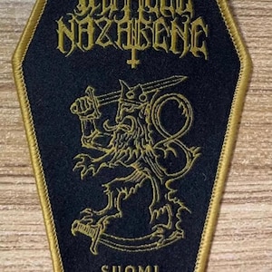 May include: Black coffin-shaped patch with gold trim featuring the band name "Impaled Nazarene" and a lion emblem. The text "Suomi Finland Perkele" is printed at the bottom. A patch for fans of metal music.