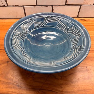 May include: A round ceramic bowl with a glossy blue glaze. The wide rim features a repeating carved pattern of white, leaf-like shapes. The center of the bowl is smooth and concave, reflecting light across its deep blue, glazed surface.