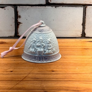 May include: A pale blue ceramic bell ornament featuring a raised relief design of three children sitting together. The bell is decorated with small star patterns and has the year 1987 embossed at the base. A light pink ribbon is attached to the top loop.
