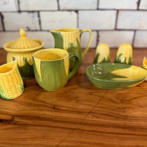 May include: A ceramic dinnerware set featuring a yellow corn kernel pattern with green husk accents. The collection includes a lidded sugar bowl, a creamer pitcher, a mug, two salt and pepper shakers, an oval dish, and a small individual bowl.