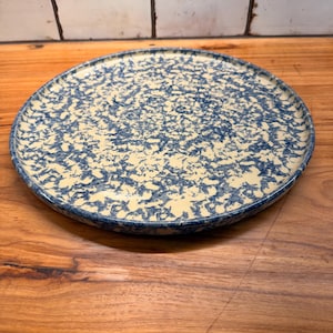 May include: A round, flat plate featuring a speckled pattern of dark blue paint splatters over a cream-coloured base. The plate has a smooth, circular rim and a shallow, wide surface area, displaying a consistent mottled texture across the entire top.