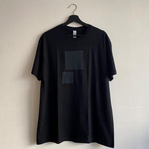 May include: Black short-sleeved t-shirt with two dark gray square patches on the front. The shirt is hanging on a black hanger against a plain, off-white wall. The t-shirt is made of a soft material.