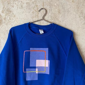 May include: A royal blue sweatshirt with a geometric design. The design features overlapping translucent squares in shades of pink and purple, outlined with orange, pink, and yellow lines. The sweatshirt is hanging on a metal hanger.