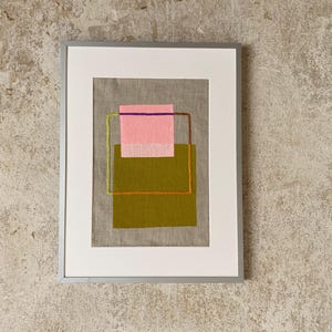 May include: Framed abstract art piece featuring a gray background with a pink and olive green geometric design. The artwork is framed in a silver metal frame with a white mat.