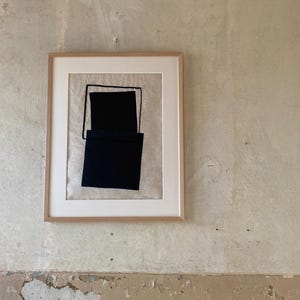 May include: Framed abstract art piece featuring a black geometric design on a neutral background. The artwork is encased in a light wood frame with a white mat. The piece is hanging on a textured, light-colored wall.