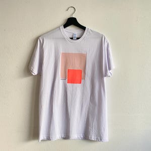 May include: White t-shirt with a geometric design. The design features a large peach-colored square over a smaller, bright orange square. The text "somewhere studio" is printed below the squares. The shirt is hanging on a black hanger.