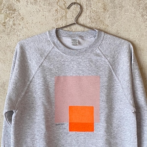 May include: A heather gray sweatshirt with a geometric design. The design features a large pink square above a smaller orange square. The text "Spiral Studio" is printed below the squares. The sweatshirt is hanging on a metal hanger.