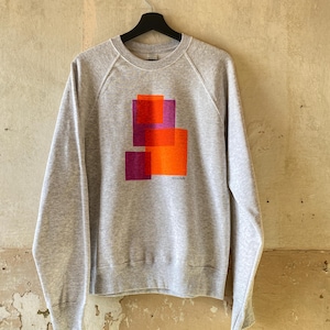 May include: A heather gray sweatshirt with a graphic design of overlapping orange and purple rectangles. The long-sleeved top is hanging on a black hanger against a neutral-toned wall.