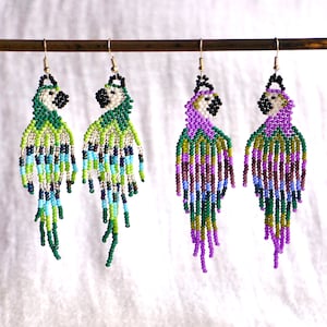 Op de afbeelding: Handgemaakte kralen papegaai oorbellen in groen, blauw en geel, en paars, groen en geel. De oorbellen hangen aan een goudkleurige draad. De papegaai oorbellen hebben een witte kop en een zwarte snavel.
