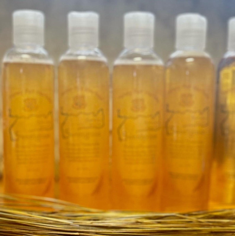 K&ouml;nnte beinhalten: Mehrere transparente Flaschen mit wei&szlig;en Verschl&uuml;ssen sind mit einer goldenen Fl&uuml;ssigkeit gef&uuml;llt. Jede Flasche hat ein Etikett mit Text und einem dekorativen Emblem. Die Flaschen stehen auf einem Bett aus goldenem Stroh.