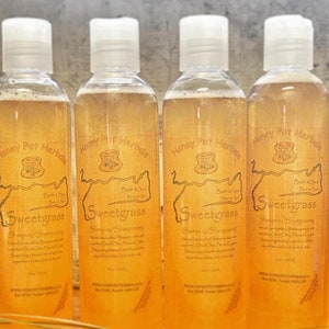 May include: Four clear bottles of Sweetgrass shampoo with white caps. The labels are tan with the text "Honey Pot Herbals" and "Sweetgrass". The shampoo is a light amber color. The bottles are lined up on a wooden surface.