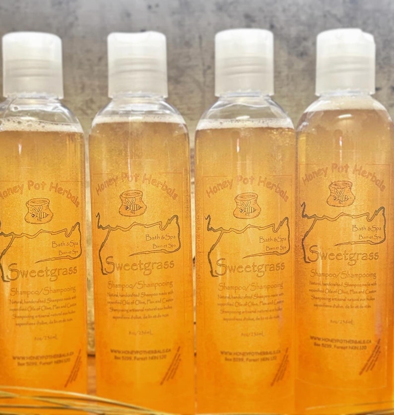 Op de afbeelding: Vier doorzichtige flessen Sweetgrass shampoo met witte doppen. De labels zijn bruin met de tekst "Honey Pot Herbals" en "Sweetgrass". De shampoo heeft een licht amberkleur. De flessen staan op een houten ondergrond.