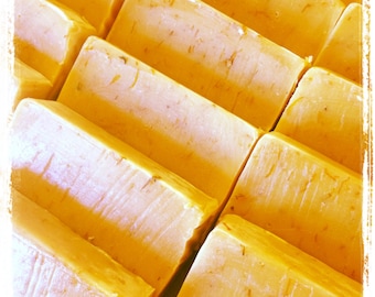 May include: Close-up of several rectangular bars of handmade soap. The soap has a light yellow color with visible flecks of what appears to be dried herbs or flowers. The bars are arranged in a diagonal pattern, creating a visually appealing display.
