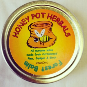 Peut inclure: Une boîte métallique ronde avec une étiquette jaune et orange. L'étiquette affiche "HONEY POT HERBALS" et une abeille de dessin animé. Le texte "Forest Balm" est également visible. La boîte contient 60 ml de produit.