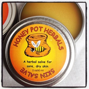 May include: A metal tin of Honey Pot Herbals skin salve. The label features a cartoon bee and the text "A herbal salve for sore, dry skin." The tin contains 2 oz / 60 ml of salve.