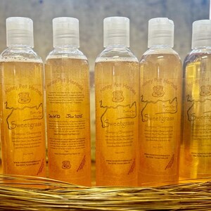 Op de afbeelding: Vijf doorzichtige plastic flessen Sweetgrass shampoo met witte klapdoppen. Elke fles heeft een etiket met tekst en een logo. De shampoo vloeistof is amberkleurig.
