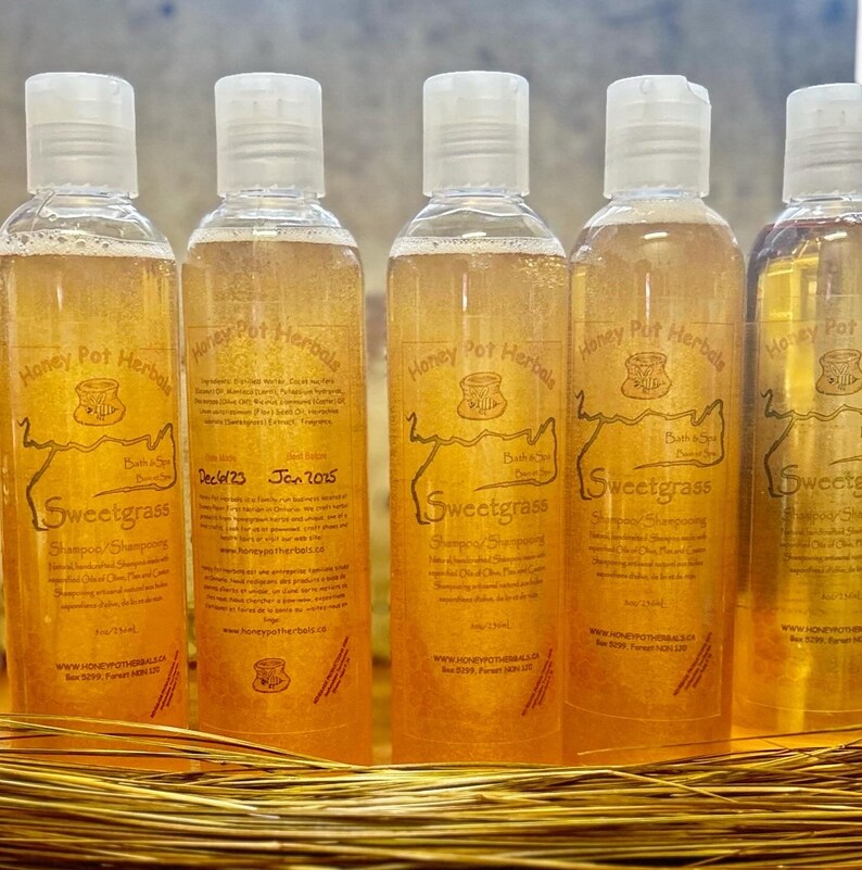 Op de afbeelding: Vijf doorzichtige plastic flessen Sweetgrass shampoo met witte klapdoppen. Elke fles heeft een etiket met tekst en een logo. De shampoo vloeistof is amberkleurig.