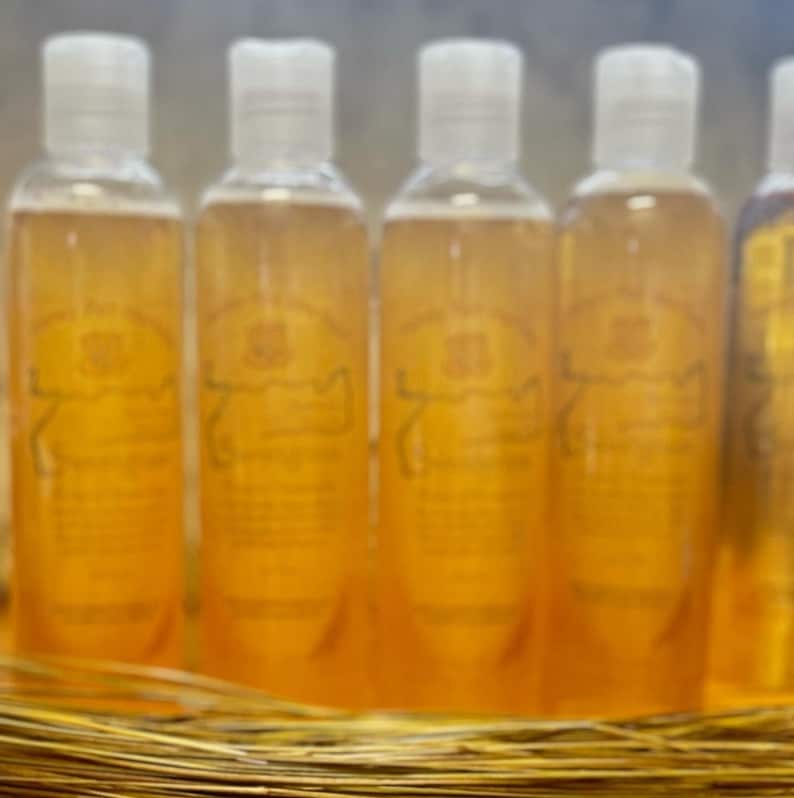 Op de afbeelding: Verschillende heldere flessen met witte doppen, gevuld met een gouden vloeistof. Elke fles heeft een etiket met tekst en een wapenontwerp. De flessen zijn gerangschikt in een rij, rustend op een bed van gouden stro.