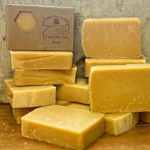Puede incluir: Una pila de barras de jabón rectangulares de color amarillo claro. Una pequeña caja de cartón marrón con una ventana hexagonal muestra una barra de jabón. La caja tiene el texto "Honey Pot Herbals" y "Touch Me Not Soap".
