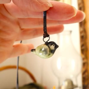 Puede incluir: Un pequeño colgante de cerámica con forma de pájaro, con cabeza negra y blanca y cuerpo verde claro, cuelga de un cordón negro. El colgante es sostenido por una mano, mostrando los detalles de la joya artesanal.