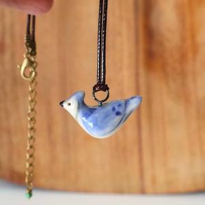 May include: A ceramic bird pendant necklace. The pendant is a small, blue bird with white accents and a brown leather cord. The necklace also includes a gold-colored chain with a clasp.