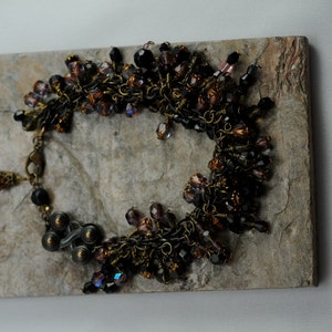 May include: A delicate bracelet made of brown wire and black, purple, and brown beads. The bracelet is arranged in a circle and has a clasp.