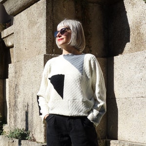 Può includere: Una donna con un maglione bianco con disegni geometrici neri, pantaloni neri e occhiali da sole si trova davanti a un muro di pietra con piante in vaso.