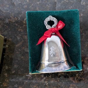 May include: A silver bell with a decorative handle and a red ribbon bow, presented in a green velvet-lined box. The bell has an embossed design on its surface, suggesting a collectible or decorative item.