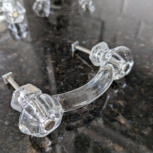 May include: A clear glass cabinet knob with a curved handle and a silver screw. The knob has a faceted design and is shown on a dark countertop.