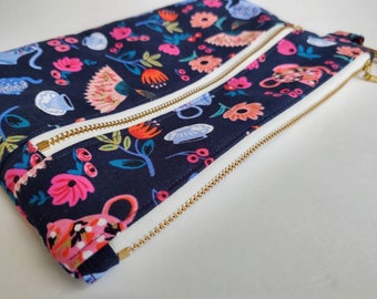 Pode incluir: Uma bolsa azul marinho com zíper, com um zíper branco e uma estampa floral com flores rosa, vermelhas e amarelas, pássaros e xícaras de chá.